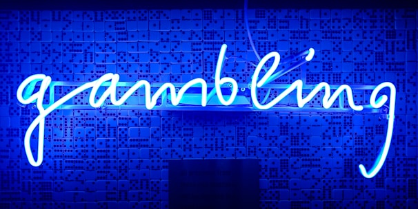 A bright blue neon sign spells out the word 'gambling' in cursive script. The background features a textured wall covered in domino tiles, also illuminated by the blue light, creating a vivid and visually striking scene.