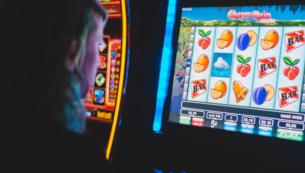A person is closely observing a digital slot machine with colorful fruit symbols, bars, and a bell. The slot machine screen displays symbols like oranges, plums, cherries, and clouds. The background is dimly lit, creating a focused and somewhat intense atmosphere.