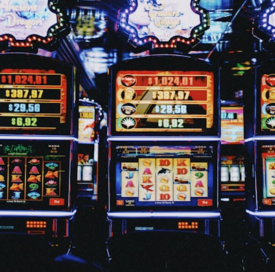 A row of colorful slot machines lit with bright neon lights, showcasing various graphical elements on the screens, indicating different games and jackpots. The atmosphere suggests a lively casino setting, bustling with excitement.