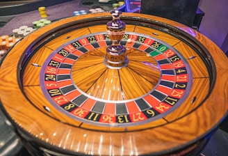 A roulette wheel with alternating red and black numbered pockets on a wooden platform. The wheel contains a small white ball resting in one of the pockets, indicating gameplay. Surrounding the wheel are stacks of colorful poker chips, suggesting a casino environment.