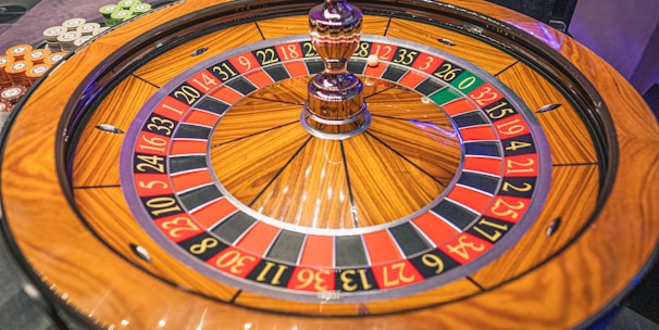 A roulette wheel with alternating red and black numbered pockets on a wooden platform. The wheel contains a small white ball resting in one of the pockets, indicating gameplay. Surrounding the wheel are stacks of colorful poker chips, suggesting a casino environment.