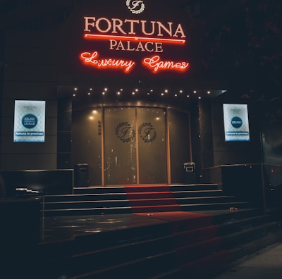 A neon-lit casino entrance with a red carpet leading up to large double doors. The sign reads 'Fortuna Palace Luxury Games' in bright red and white neon lights. The dimly lit atmosphere suggests a night-time setting. There are two digital displays on either side of the entrance showing promotional content.