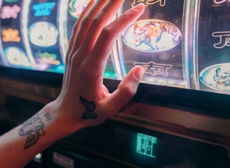 A hand with tattoos reaches towards a brightly lit slot machine, featuring various colorful symbols including a tiger. The image captures the moment of anticipation as the person engages with the machine during a game of chance.