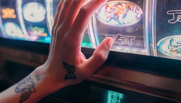 A hand with tattoos reaches towards a brightly lit slot machine, featuring various colorful symbols including a tiger. The image captures the moment of anticipation as the person engages with the machine during a game of chance.